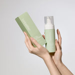 Hands holding a green skincare product and packaging against a light gray background